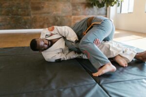 Two men demonstrate Brazilian Jiu-Jitsu grappling techniques indoors on a gym mat.